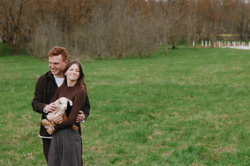 A joyful couple outdoors in a green field cuddle a goat kid, embodying rural lifestyle and bonding with animals. A tender moment of connection and simple countryside living.
