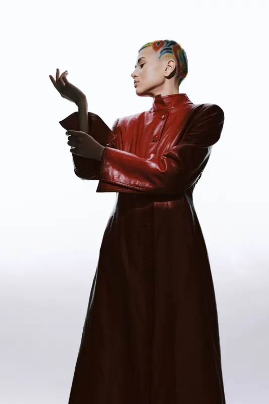 Stylish woman with colorful braided hair wearing a long red leather coat, posing confidently against a simple white background in a modern fashion portrait.