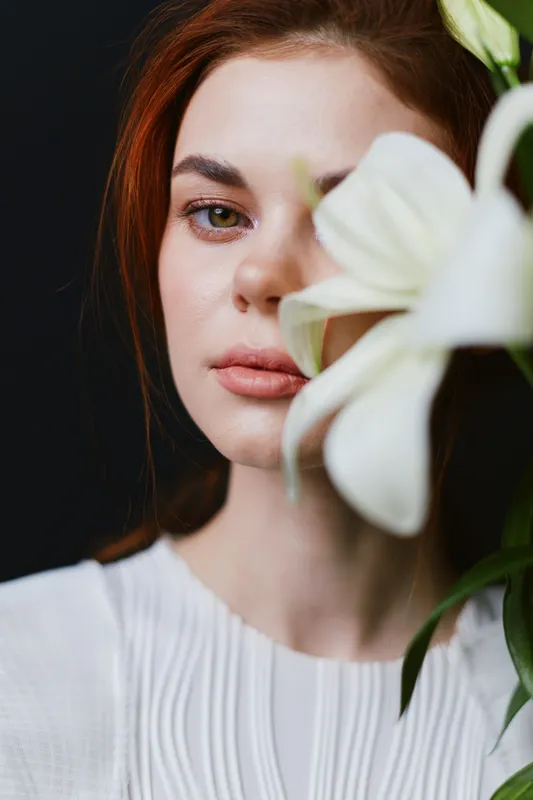 Serene portrait of a young woman with red hair, softly gazing at the camera, adorned with a white flower against a dark background, emphasizing natural beauty and elegance