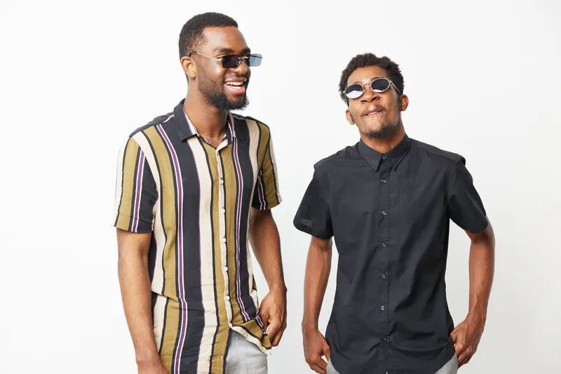 Two cheerful African American men wearing stylish sunglasses and casual shirts, sharing a joyful moment together, showcasing happiness and camaraderie in a bright environment.