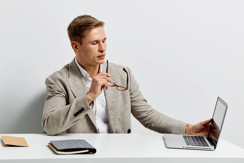 Professional man wearing formal blazer and white shirt working on a laptop in a modern office environment holding eyeglasses. Business, technology, workspace, entrepreneur, communication, corporate co