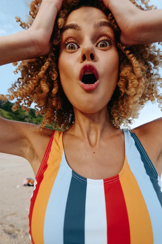 Young woman at the beach wears a colorful striped swimsuit and has a surprised expression as she raises her hands to her head, bright sky overhead and a carefree summer mood.
