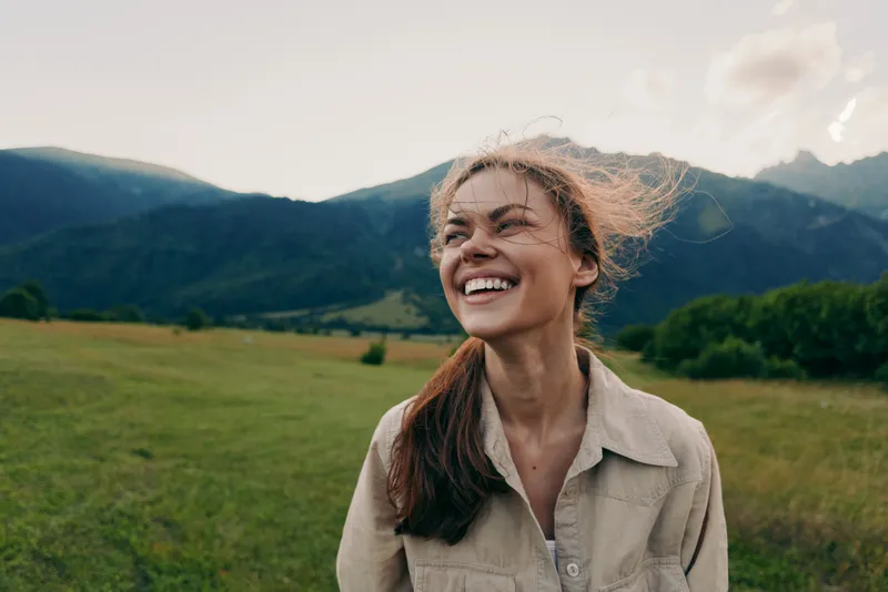 Joyful outdoor lifestyle portrait of a woman in a field near mountains, warm sunlight, casual attire, natural smile, candid moment of happiness outdoors and light breeze.