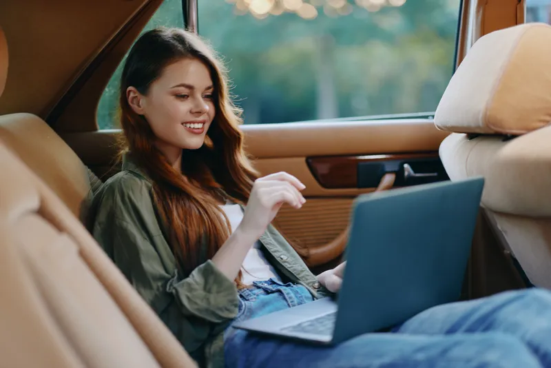 Young woman using a laptop in a car, wearing casual clothing with a cheerful expression, surrounded by nature. Perfect for concepts on remote work and technology usage.