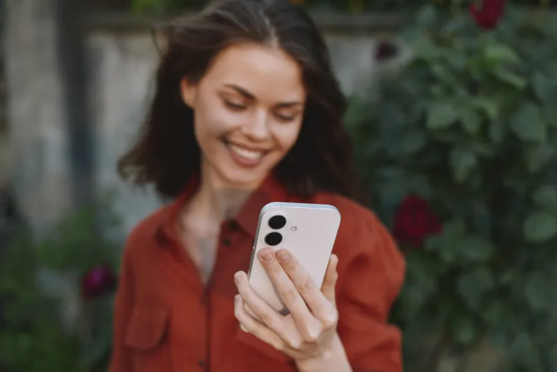 Smiling woman in a casual red shirt holding a smartphone outdoors, with natural light and a lush garden background, conveying lifestyle, wellness, and modern connectivity and daily inspiration