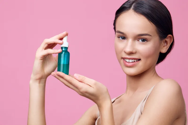 Young woman holding a green cosmetic bottle against a pink background, showcasing healthy skin and beauty products in a vibrant and cheerful setting