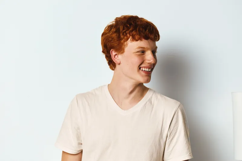 Young man with curly red hair wearing a white t shirt standing against white wall smiling and looking to side in casual style with natural light indoors.