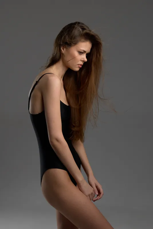 young woman in black bodysuit posing with serious expression against gray background