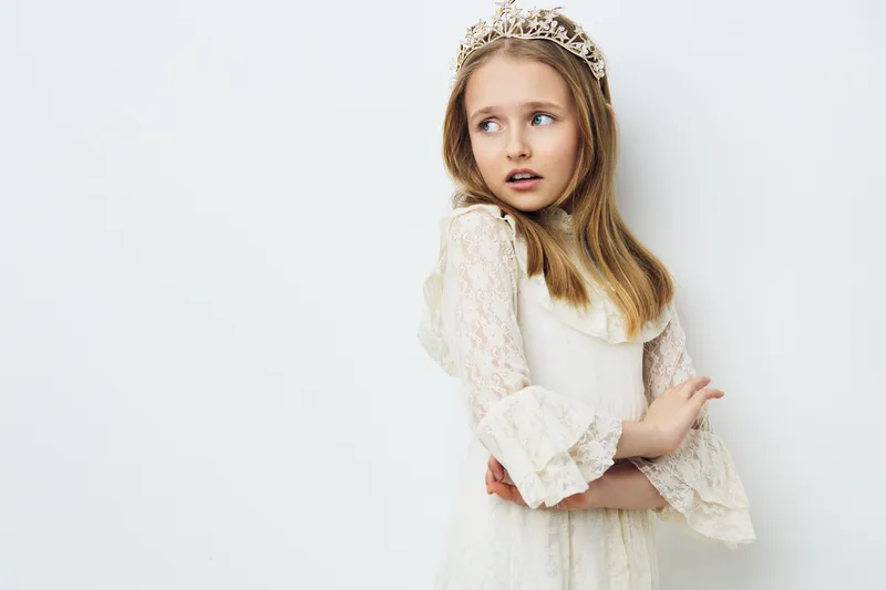 A young girl wearing a white lace dress and tiara, expressing a thoughtful emotion against a soft white background, ideal for concepts of innocence and childhood dreams