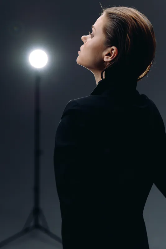 Profile of a woman with blonde hair in black clothing looking up with studio light in the background, minimalistic dark setting.