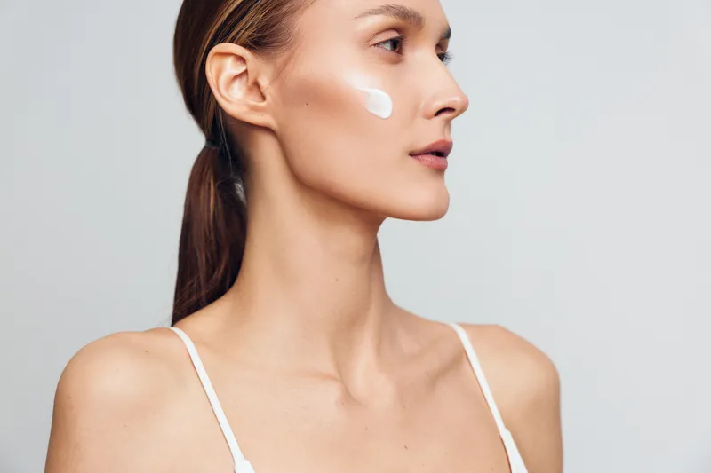 Young woman with long brunette hair applies white cream on her cheek while looking sideways against a neutral background in a skincare beauty routine concept.