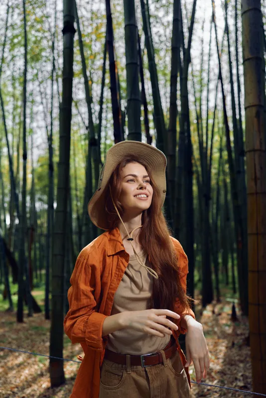 Woman in a straw hat smiles in a bamboo forest, outdoor portrait in nature with long red hair, casual autumn outfit and relaxed pose among tall green stalks and soft light.