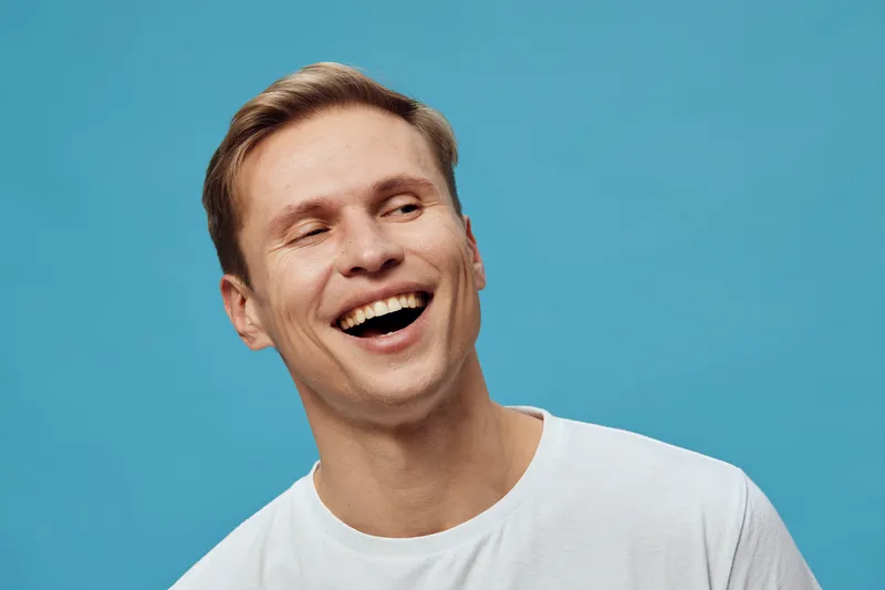 Smiling young man with fair skin and short brown hair wearing a white t-shirt against a bright blue background, showing happiness and joy. Perfect for lifestyle, positive emotion, human expression, ca