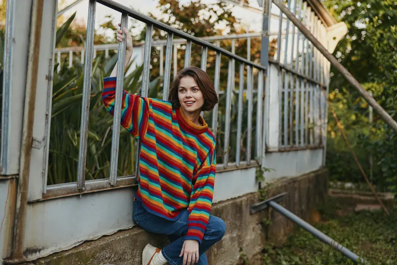 Woman in rainbow sweater smiling and posing at greenhouse window, casual portrait outdoors with plants and vintage glass, cozy knitwear and jeans sitting on ledge holding railing.