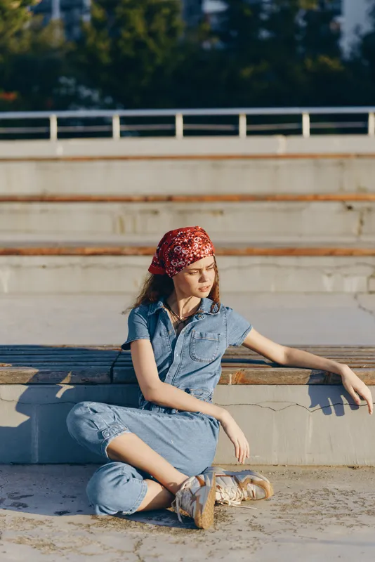Lifestyle woman in modern boho-western denim street style sitting outdoors, wearing red bandana and casual shoes under warm natural sunlight, film color mood.