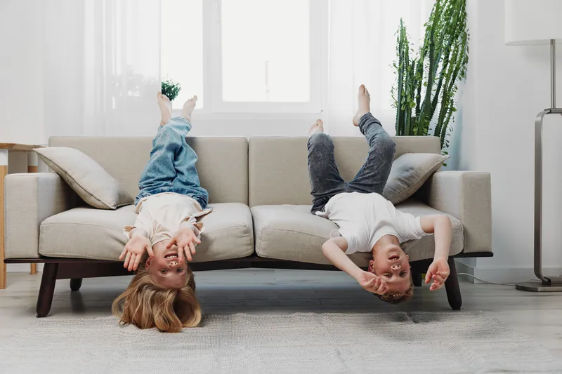 Two playful children hanging upside down on a beige sofa in a bright living room with natural light and indoor plants.