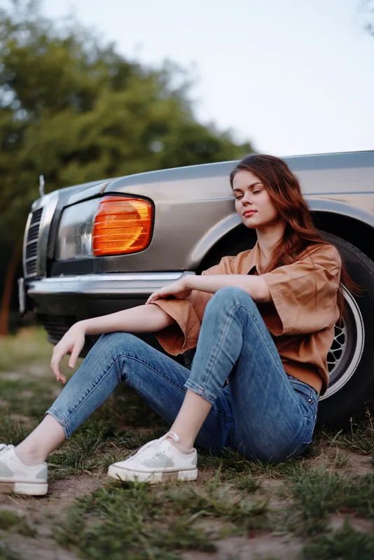 Young woman sitting beside a vintage car, wearing casual attire, embodying a relaxed and serene mood in a nature setting.
