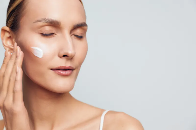 A young woman with clear skin applies cream to her face with closed eyes in a minimalistic setting. Concept for skincare, beauty, self-care, and healthy lifestyle.