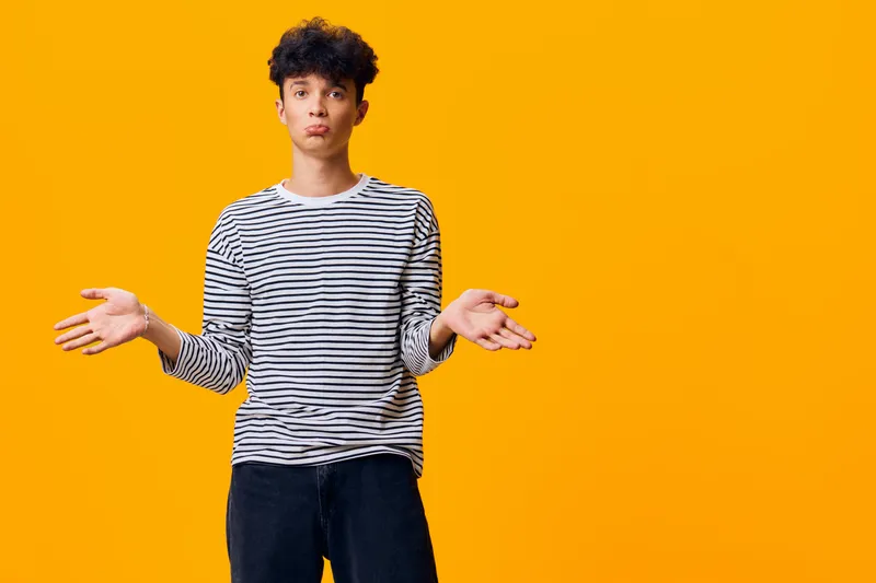 young man with curly hair wearing striped shirt stands confused with hands raised against bright orange background, casual style and puzzled expression