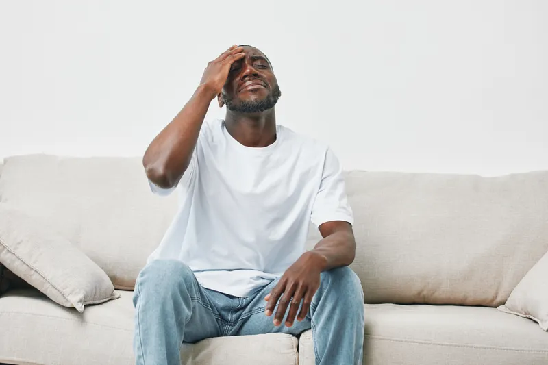 Upset young African man sitting on a sofa, holding his head in distress, expressing feelings of frustration or anxiety, wearing a casual white t shirt and jeans