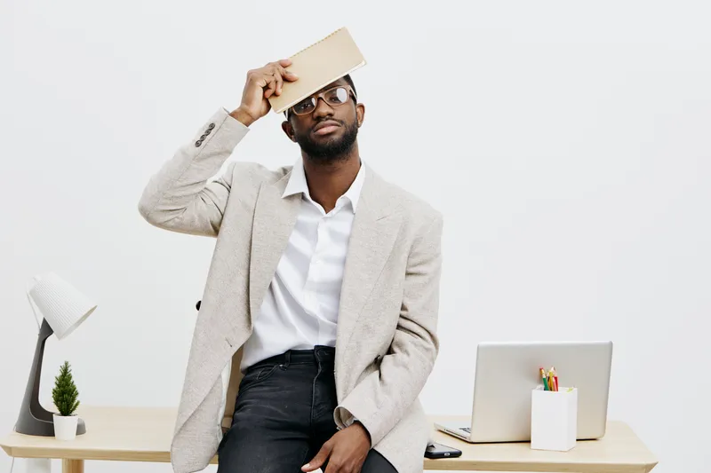 Confused black man sitting at modern desk in office holding tablet above head, expressing frustration or questioning, with laptop and plant on background, indoor shot, professional environment, expres
