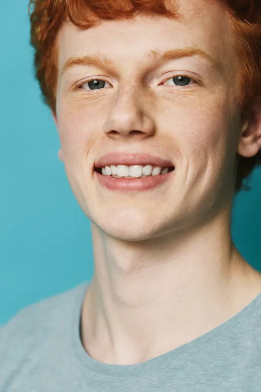 Young man with red hair and freckles smiling closeup with blue background in casual shirt showing teeth confidence and happiness in portrait photography.
