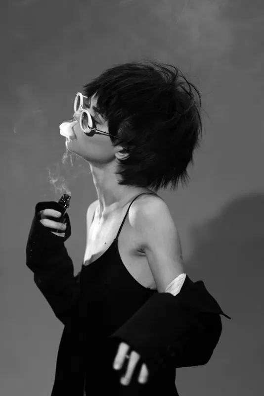 Black and white portrait of a young woman smoking with retro sunglasses and messy hair in moody lighting.