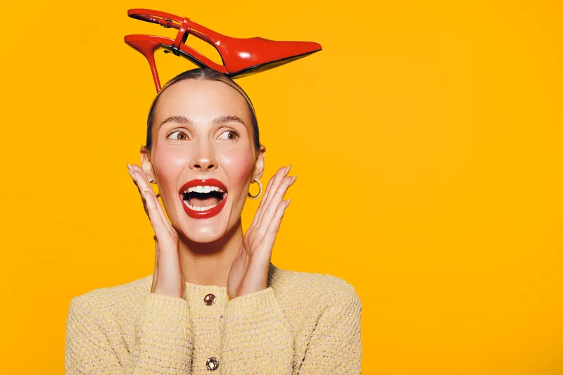 Happy woman with red high heel shoes on her head smiles excitedly on bright orange background. Concept of fashion, fun, creativity, and joyful expression in studio.