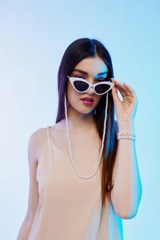 Young woman beauty fashion model wearing stylish white sunglasses and beige top poses with long straight hair in soft studio lighting on a light background. Concept for style.