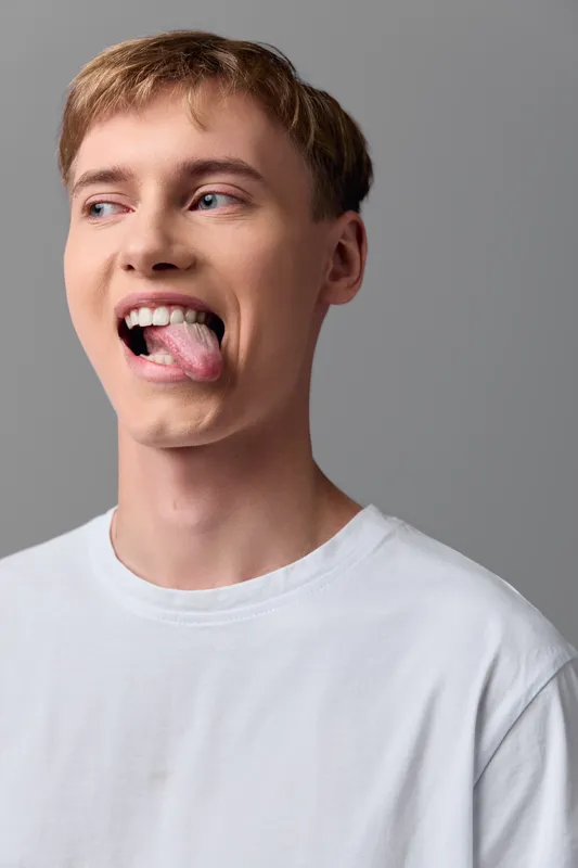 Young man with a playful tongue out expression wearing a white shirt, casual look and relaxed style in a minimal studio setting, capturing a candid, energetic moment for lifestyle or youth fashion use