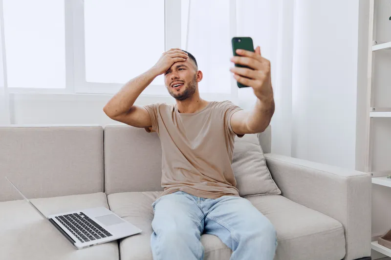 Smiling man sitting on a couch, excitedly video calling using a smartphone. Bright, modern living room with a laptop on the side adds to the cheerful atmosphere.