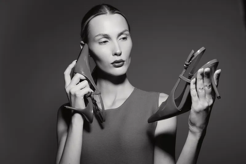 Portrait of a fair skinned Caucasian woman with slicked back hair holding two high heel shoes, one near her face and one in hand, isolated on dark background. Concept of fashion choice.