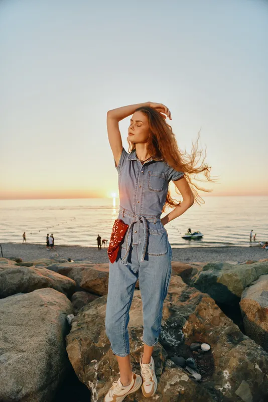 Lifestyle woman in modern boho-western denim street style posing confidently at sunset by the rocky beach with film color tones and relaxed natural mood.