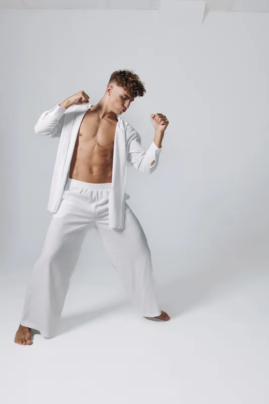 Young man with bare chest wearing white loose pants and open shirt posing in martial arts stance on white background. Fitness, strength and balance concept with confident expression.