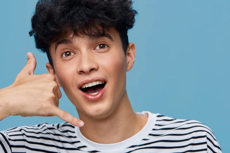 smiling man with curly hair making call me gesture wearing striped shirt on blue background, expressing friendly and playful emotion indoors.