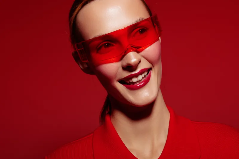 Smiling woman wearing red sunglasses and red outfit poses against red background. Fashion portrait with vibrant colors and confident expression. Concept of style and modern look.