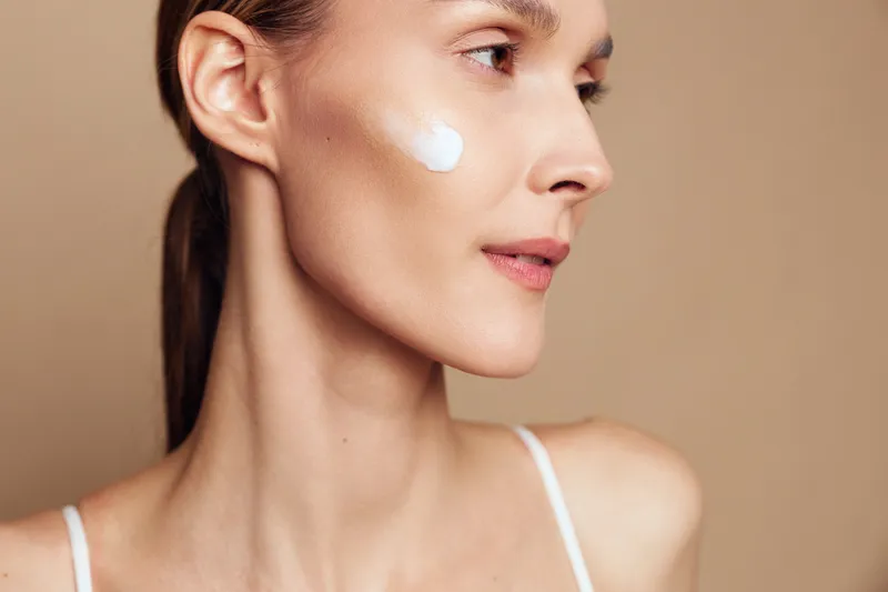Young woman with clean skin applies moisturizing cream on cheek shows natural beauty and skincare routine isolated on beige background with soft lighting and gentle expression
