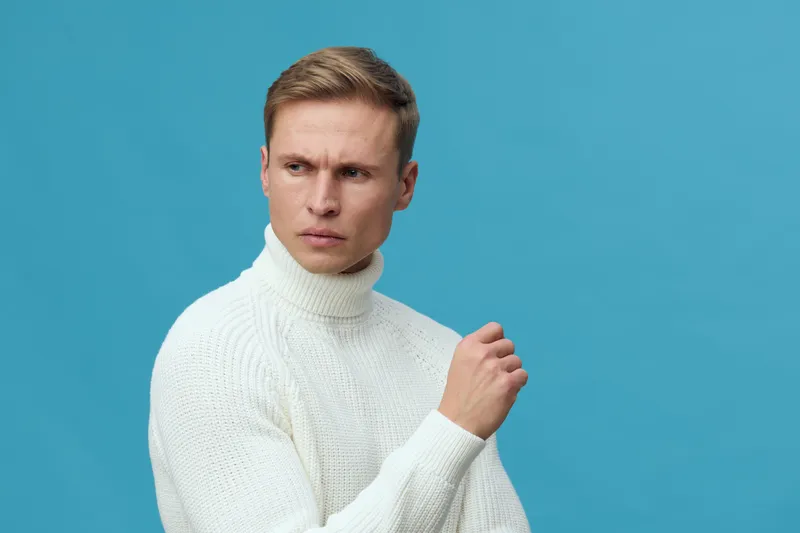 serious young caucasian man wear white knit turtleneck sweater look away thoughtful pose in studio against blue background mental health and gadgets concept