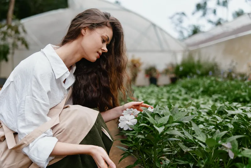 Young woman in a greenhouse, gently touching flowers, wearing a light shirt and apron, surrounded by lush greens, showcasing tranquility and connection with nature