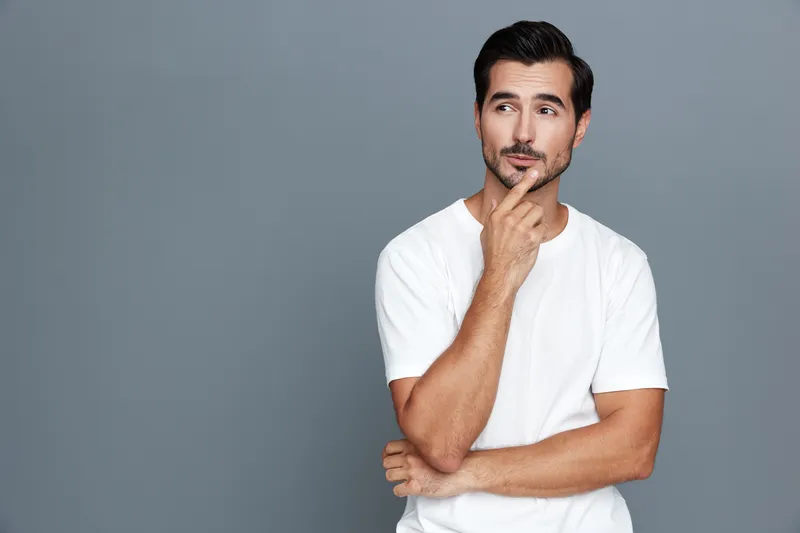 Thoughtful handsome young man wearing a casual white t-shirt posing against a solid grey background, appearing contemplative and confident in studio portrait.