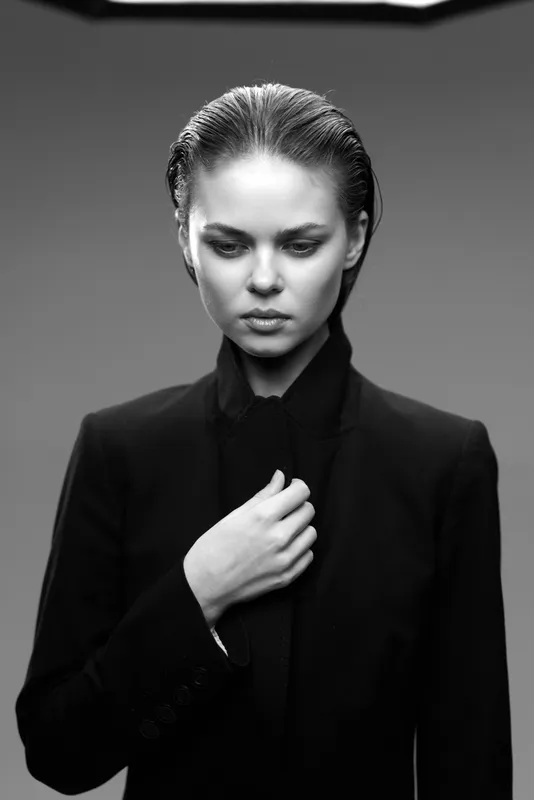 Pensive woman in black suit with slicked-back hair in moody black and white portrait expressing contemplation and elegance.
