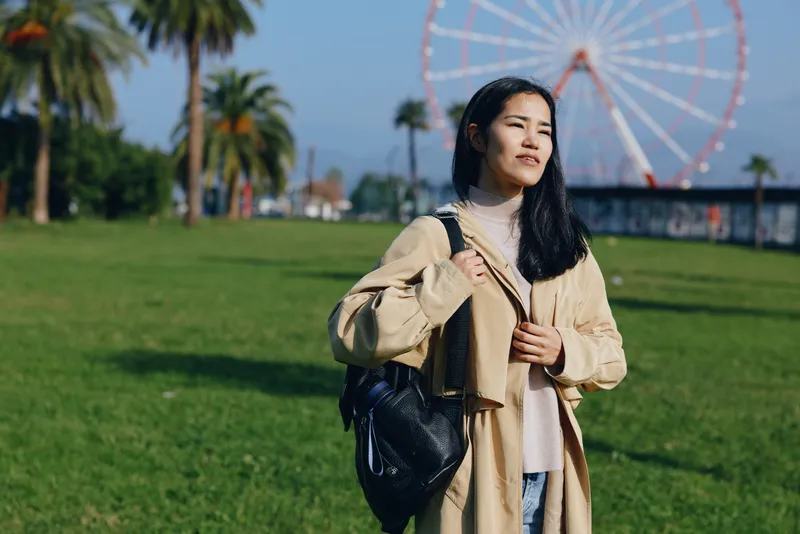 Woman in an outdoor park wearing a beige coat and bag beside a ferris wheel, portraying travel fashion and leisure under bright sunlight and blue sky