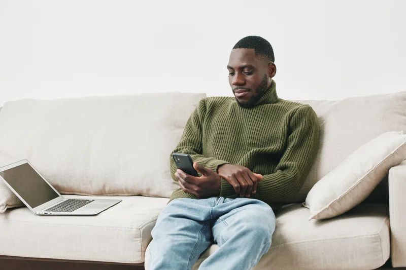 Thoughtful African American man in green sweater using smartphone while sitting on a beige sofa, emphasizing modern lifestyle and technology
