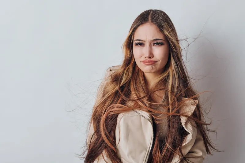 Young woman with long hair making funny face expression in casual jacket standing against light wall indoors. Emotional portrait of female with playful mood and natural look.