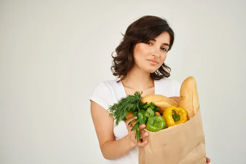 Весёлая женщина с пакетом продуктов в супермаркете на светлом фоне
