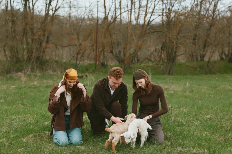Cheerful outdoors scene with three adults bonding with a playful goat kid, sunlit meadow setting, casual attire, candid interaction, sense of warmth, natural hues, rustic charm and joy