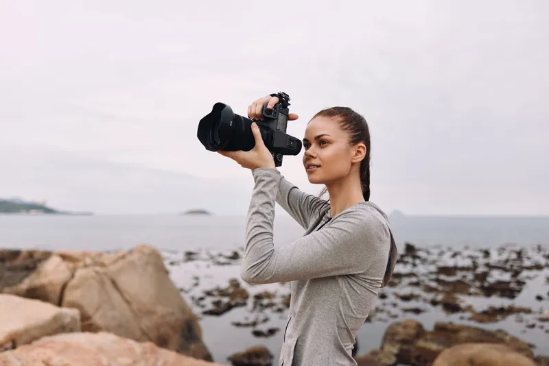 Young woman photographing scenic coastline, wearing a gray long sleeve shirt, demonstrating passion for photography on a cloudy day at the beach