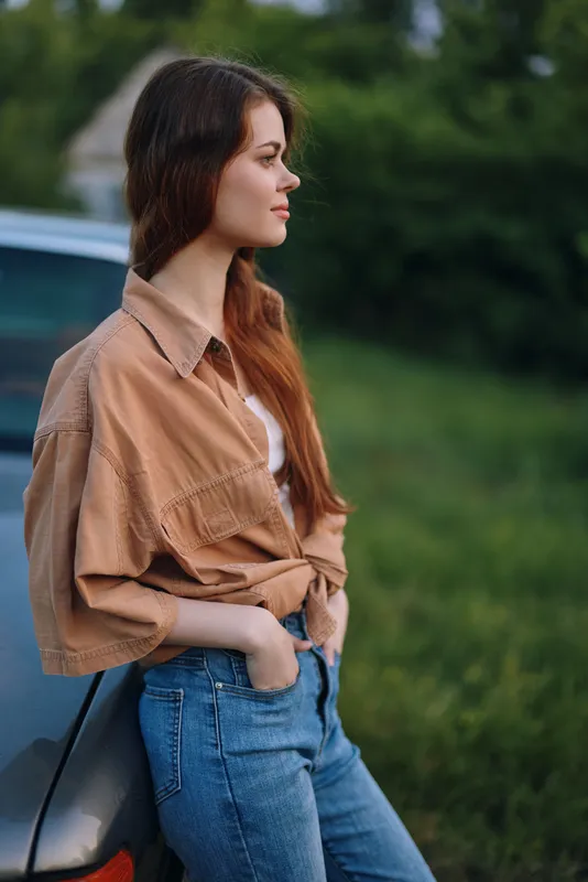Young woman in casual outfit leaning against a car in a green field, showcasing natural beauty and serenity in a summer setting.