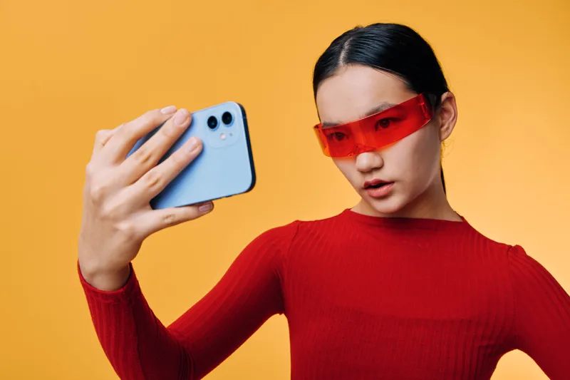 Young woman wearing red glasses taking selfie with smartphone on orange background. Female portrait with casual style for social media and digital photography content creation.