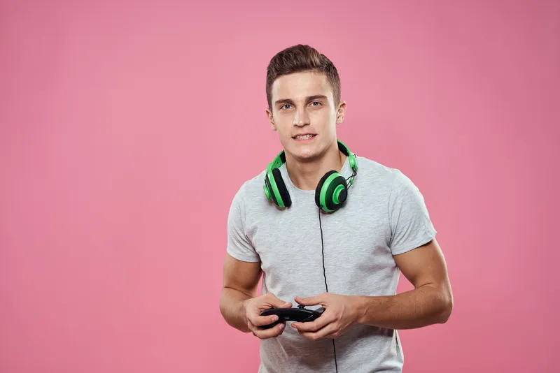 Мужчина с джойстиком в руках и в наушниках, играет в игры, развлечения, образ жизни, белая футболка, розовый фон